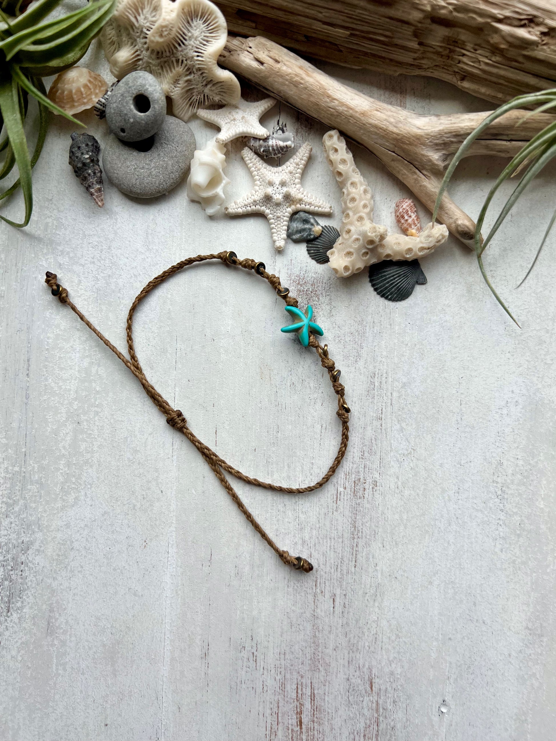 A blue starfish anklet laying on a white background surrounded by sea shells and driftwood that was found on the beaches of Florida