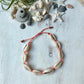 a cowrie shell anklet laying on a white background with sea shells, starfish and driftwood surrounding it. the beach inspired anklet has 9 cowrie shells on coral colored waxed cord