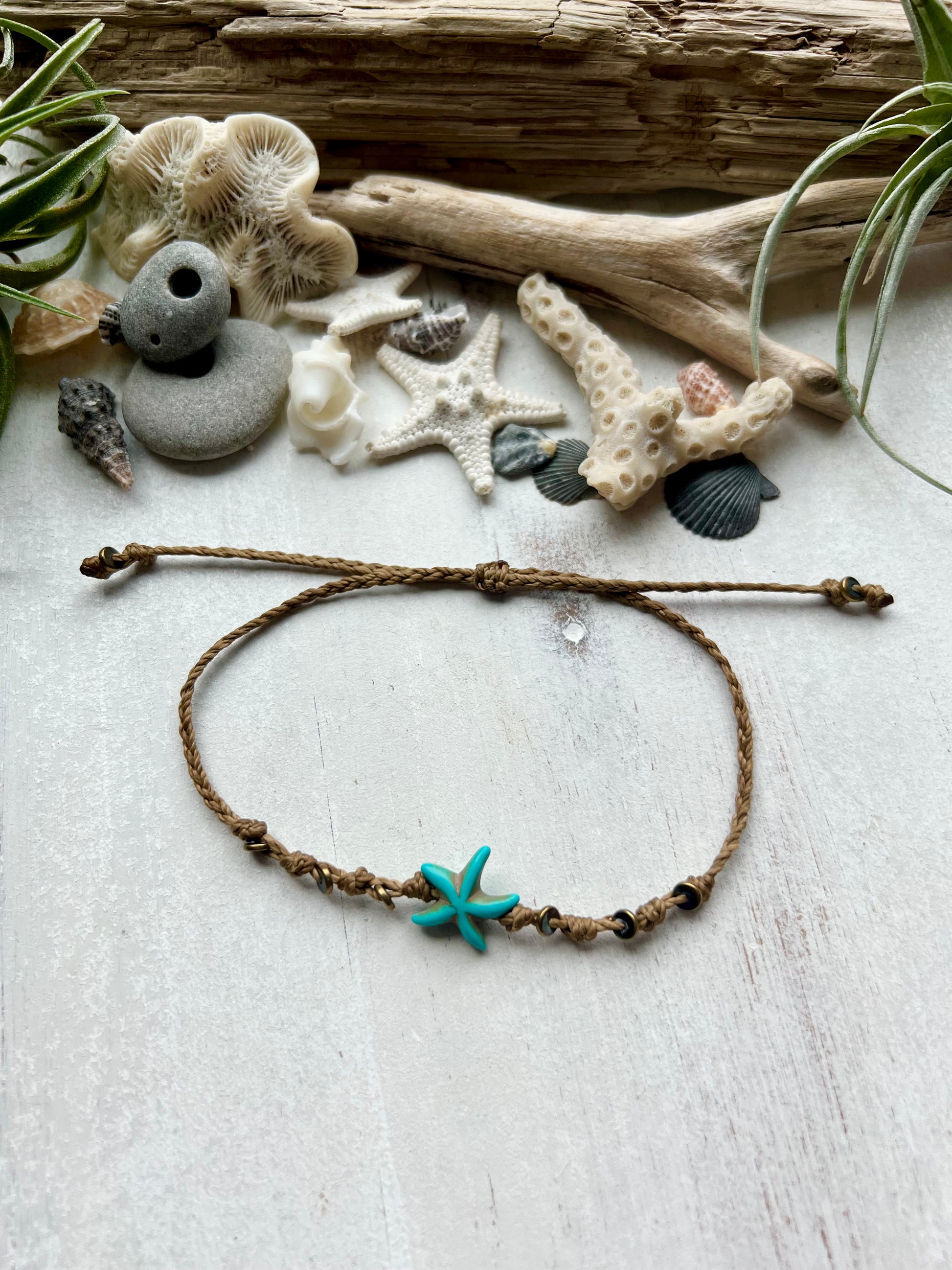 A blue starfish anklet laying on a white background surrounded by sea shells and driftwood that was found on the beaches of Florida