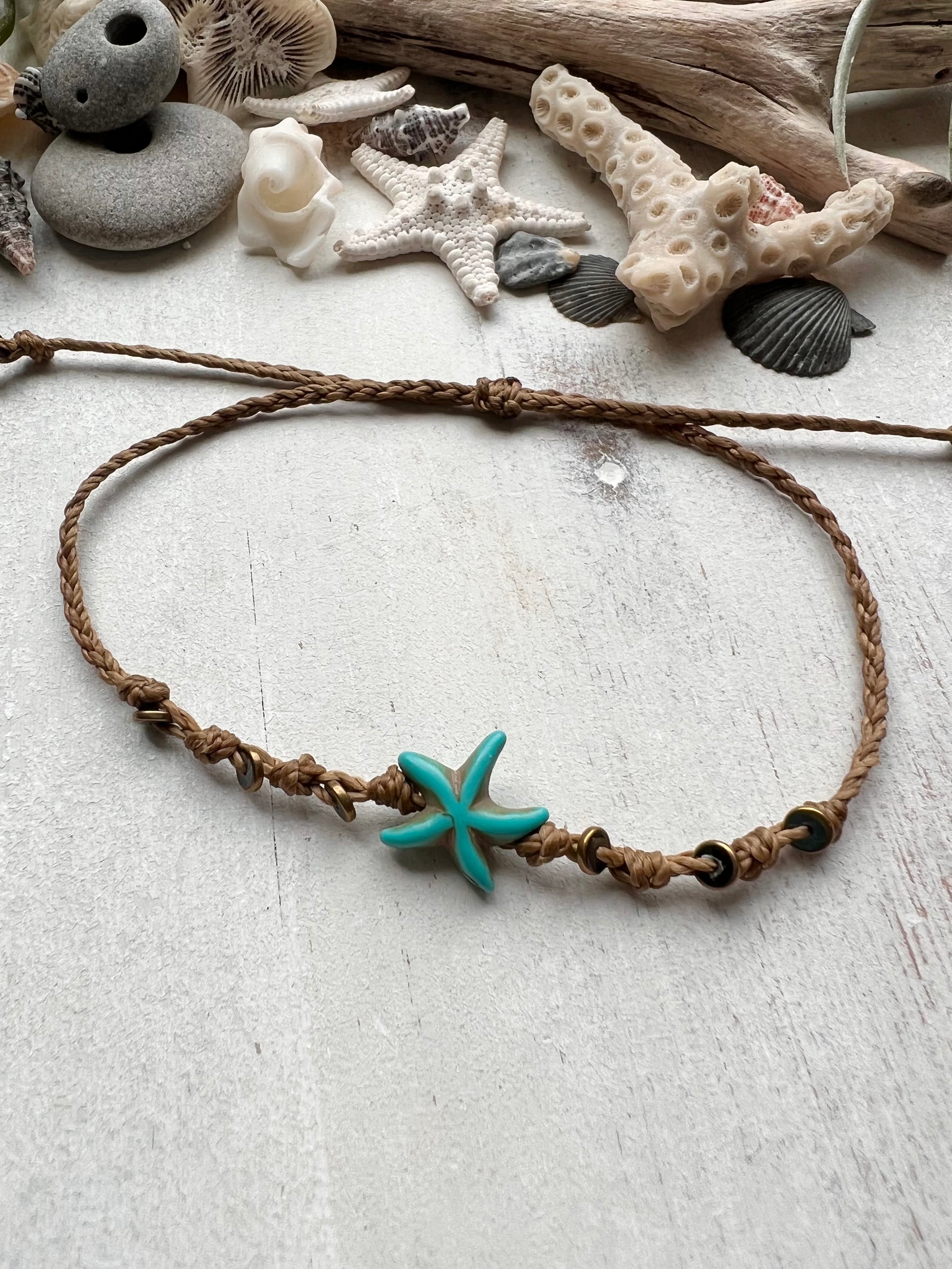A blue starfish anklet laying on a white background surrounded by sea shells and driftwood that was found on the beaches of Florida