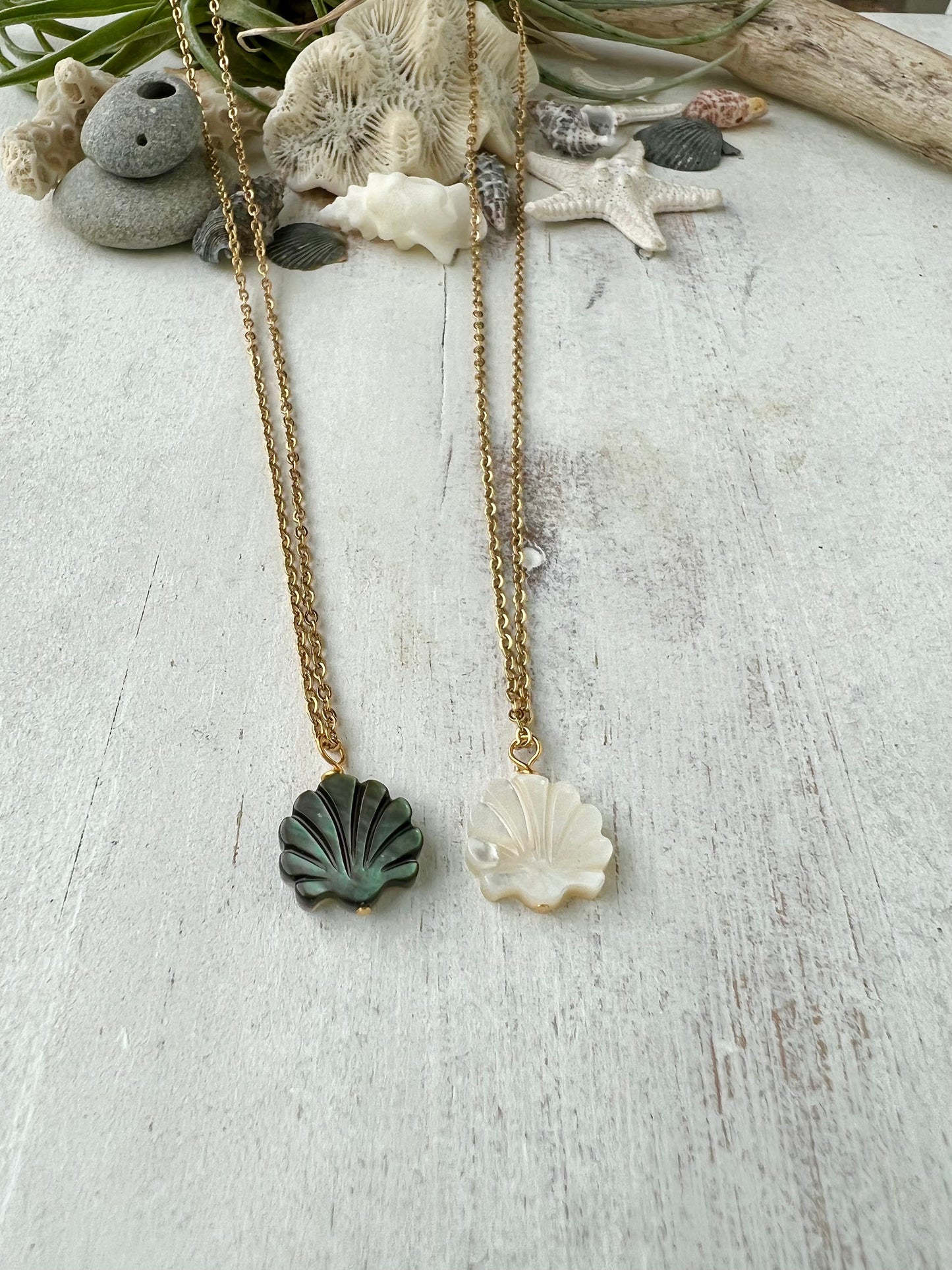 two necklaces laying on a white background with shells and coral in the background. the necklaces feature scallop shell charms in mother of pearl and black lip shell