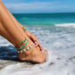 Womens feet on the beach with the ocean waves touching her toes. She is wearing three waterproof anklets, one anklet features a blue starfish charm on brown waxed polyester cord. One features heishi vinyl disc beads in turquoise. The 3rd beachy anklet features real cowrie shells, on black waxed cord. True mermaid vibes