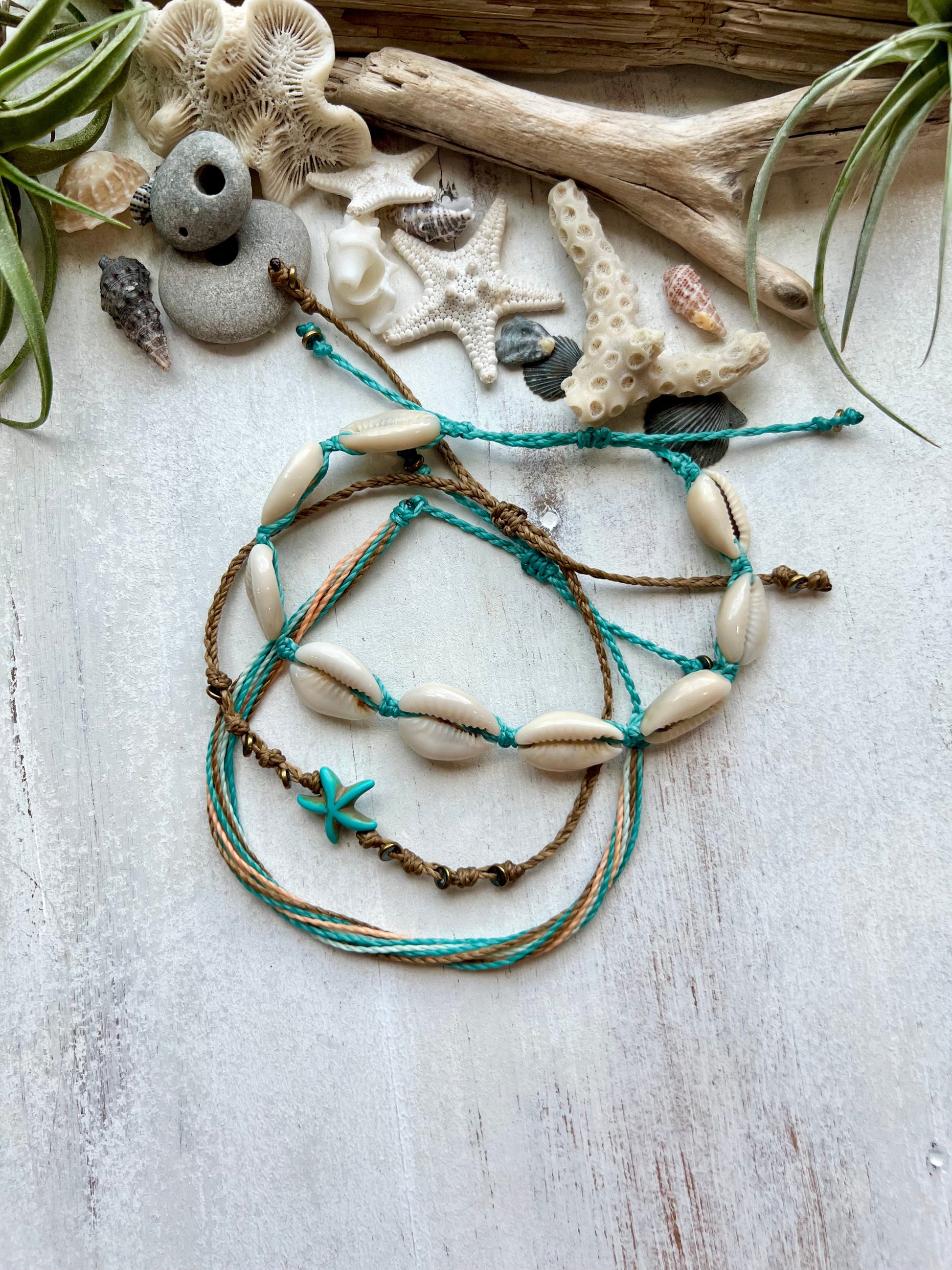 Three beachy anklets lying on a white background surrounded by sea shells, starfish, and driftwood. One anklet is multiple strands of waxed cord in shades of mint, aqua, coral and brown. One is a blue starfish charm on brown waxed cord. The third anklet has natural cowrie shells all the way around on dark mint waxed polyester string. The anklets are adjustable with sliding lock closures. 