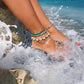 Womens feet hanging off of rocks on the beach with the ocean crashing on her feet. She is wearing Three beach inspired anklets. one anklet features a blue starfish charm on brown waxed polyester cord. One features heishi vinyl disc beads in turquoise. The 3rd beachy anklet features real cowrie shells, on black waxed cord. True mermaid vibes